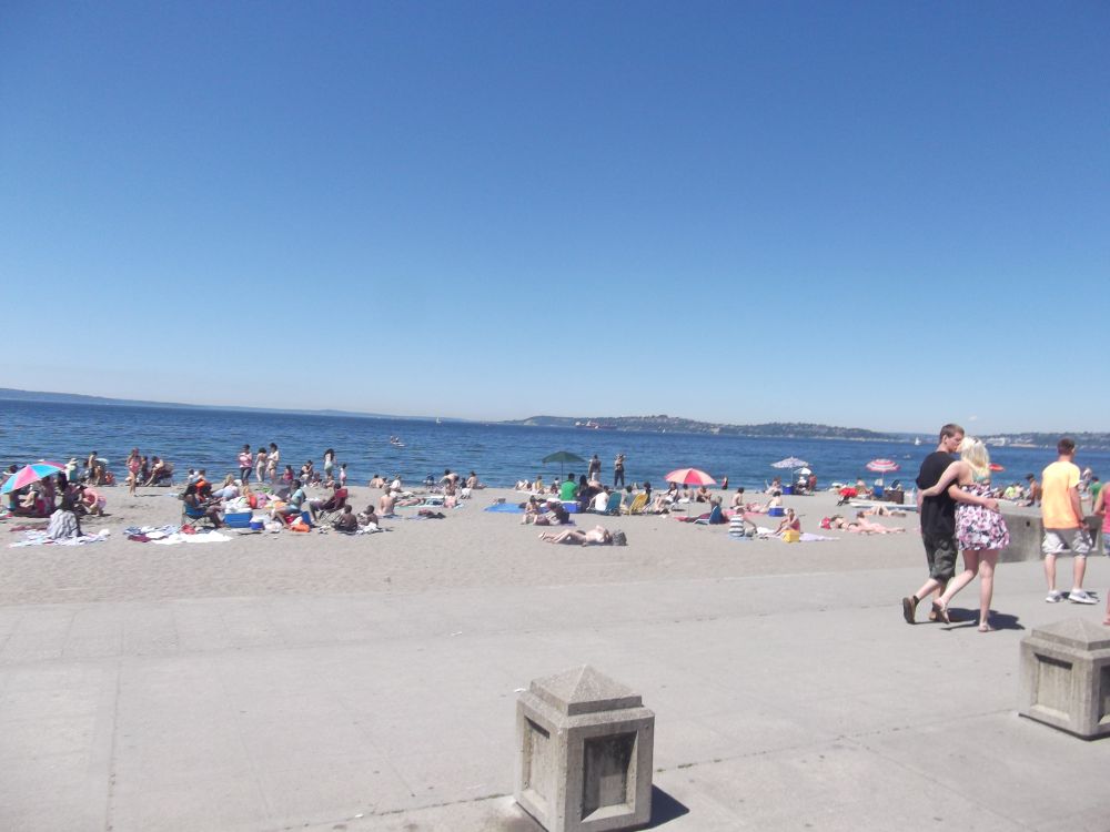 Alki Beach in June with visitors enjoying the warm summer sunshine on the sandy shoreline, the Seattle skyline reflected faintly in Elliott Bay behind them